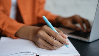 A person using both a paper notebook and pen alongside a laptop computer